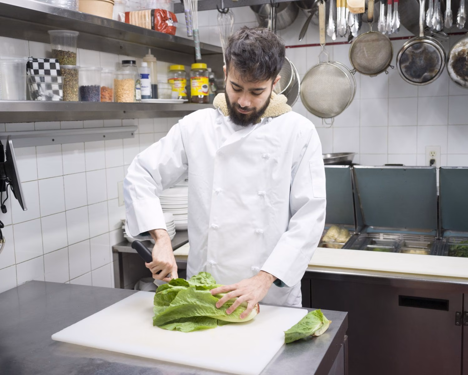 Chef in white coat chops romaine lettuce on a cutting board in a commercial kitchen.