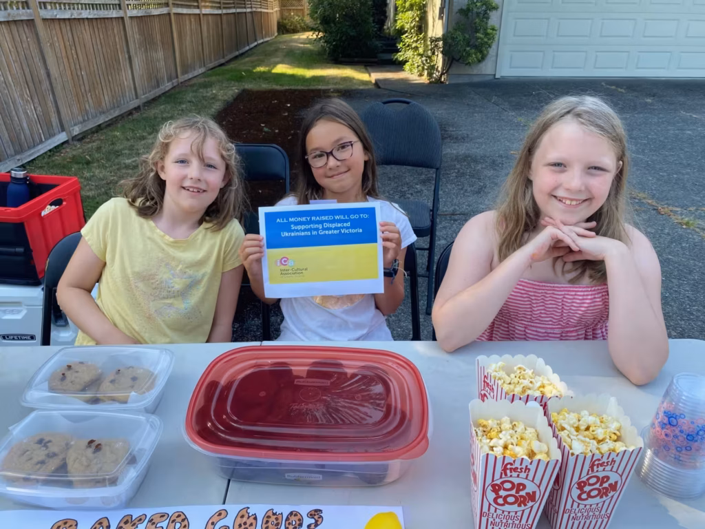 Three smiling girls at a table with cookies and popcorn, holding a sign for charity.