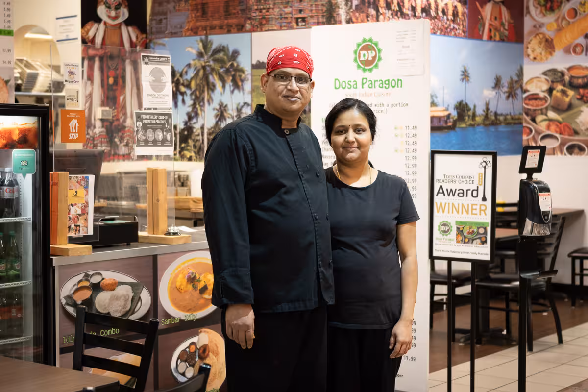 A man in a black chef's coat and red bandana stands with a woman in a black shirt, in front of a restaurant mural.