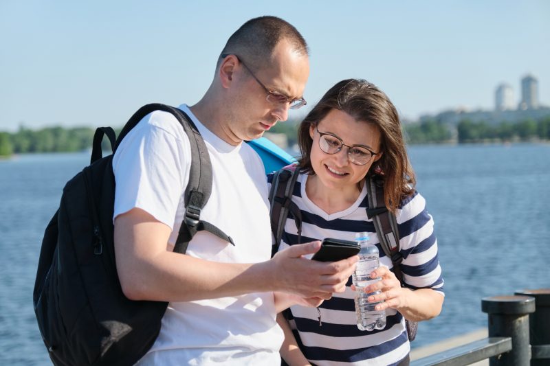Man Woman Backpacks Look Smartphone Together Body Water A man and woman with backpacks look at a smartphone together by a body of water.