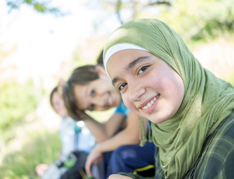 Three Children Two Boys Girl Wearing Green Hijab Three children, two boys and a girl wearing a green hijab, smile at the camera outdoors.