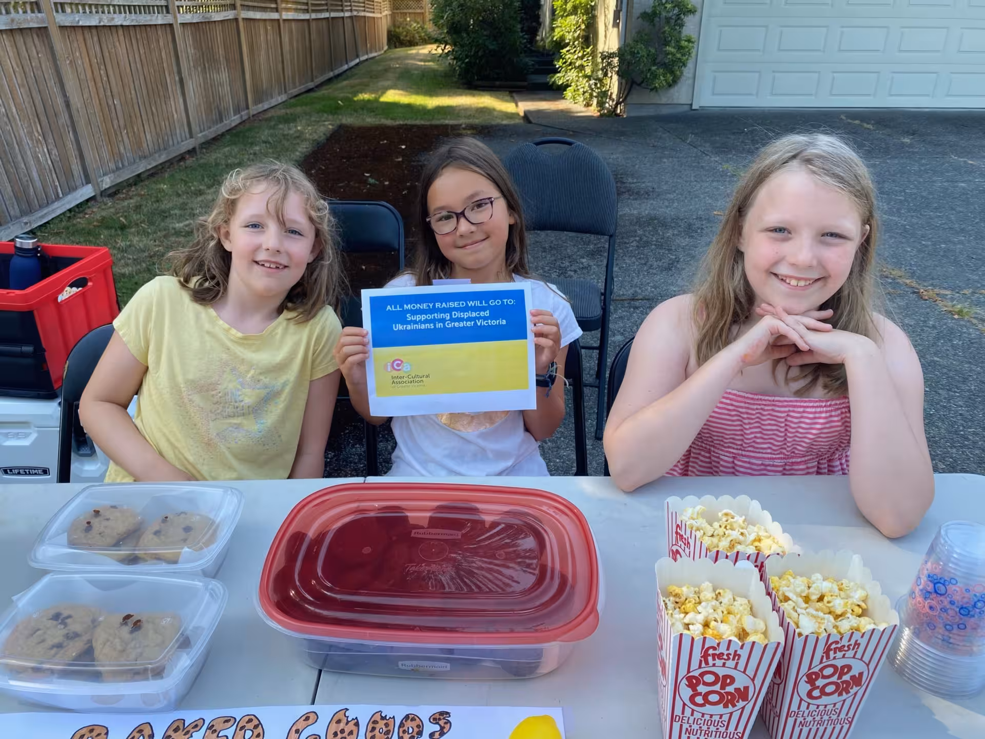 Three smiling girls at a table with cookies and popcorn, holding a sign for charity.