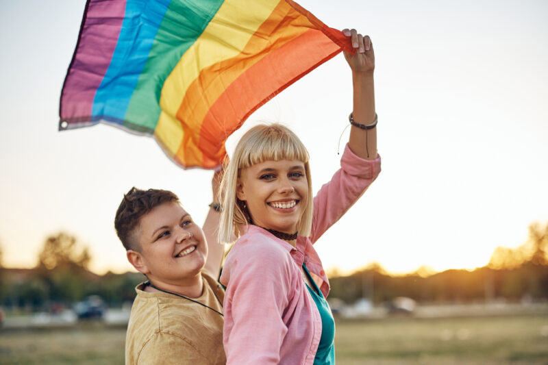 Two Smiling People Hold Rainbow Flag Aloft Field Two smiling people hold a rainbow flag aloft in a field during sunset.