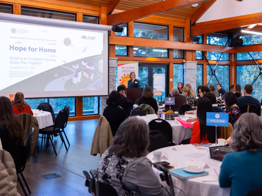 A speaker presents to a seated audience at tables in a room with large windows.