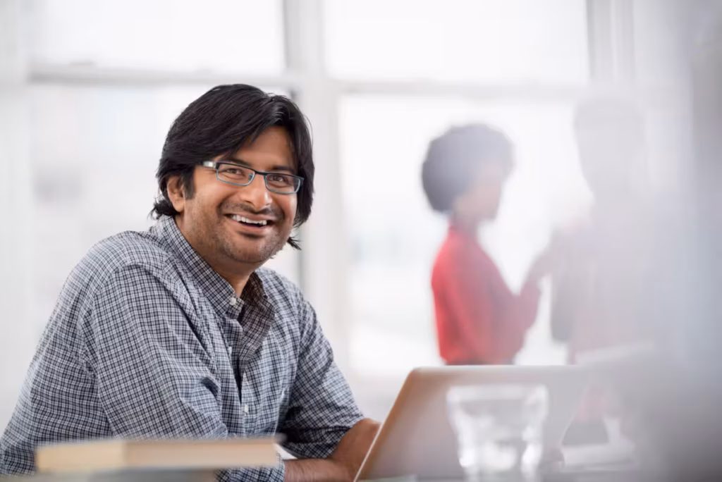 A man wearing glasses and a plaid shirt smiles at the camera, seated at a desk with a laptop.