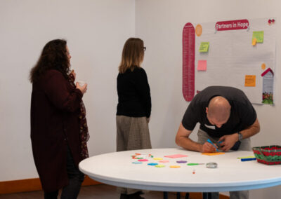 Three people stand near a whiteboard with sticky notes, one man writes on a card at a table.