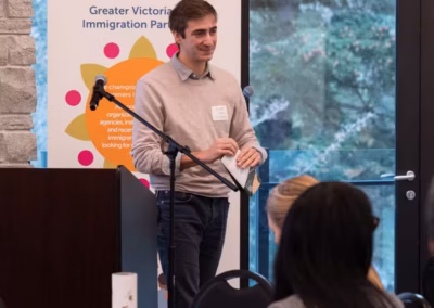 Man in light-colored sweater and jeans stands at a podium with a microphone, speaking to an audience.