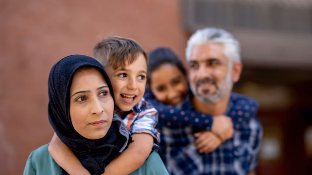 A Muslim mother wearing a hijab carries her son on her back while her husband and daughter are blurred in the background.