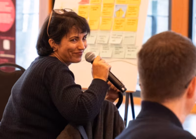 A woman in a dark sweater holds a microphone while looking back over her shoulder at a meeting.