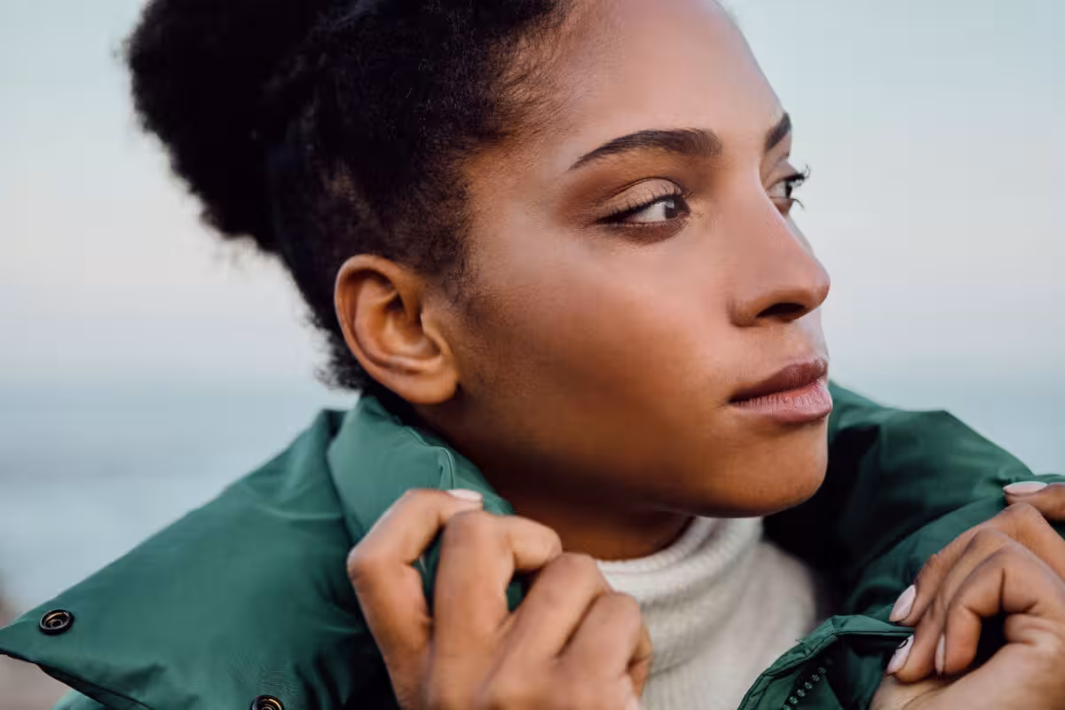 Close-up profile of a Black woman wearing a green puffer jacket and white turtleneck, looking to the right.
