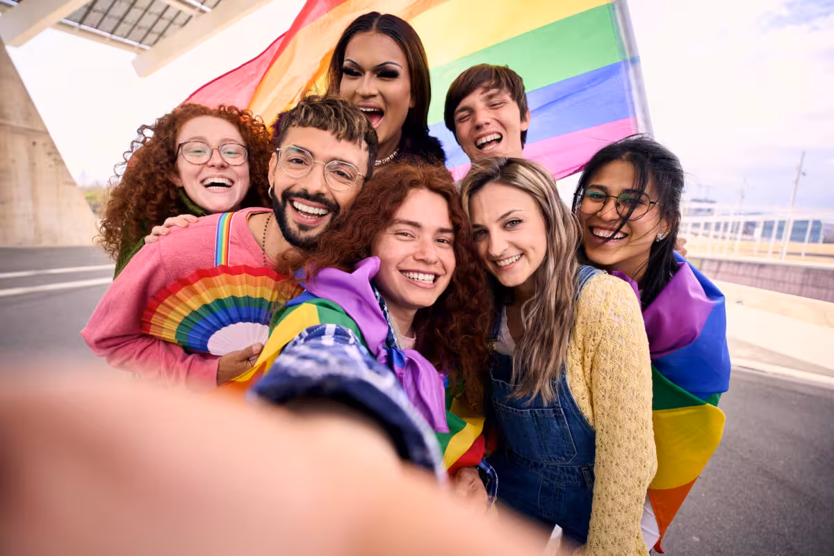 A diverse group of seven smiling people pose for a selfie with a rainbow flag and fan, celebrating Pride.