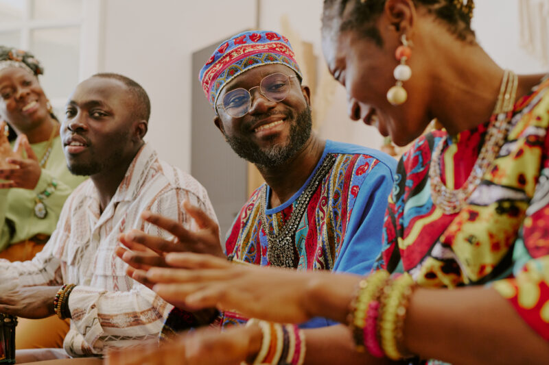 A group of Black people in colorful, traditional clothing are smiling and clapping their hands together.