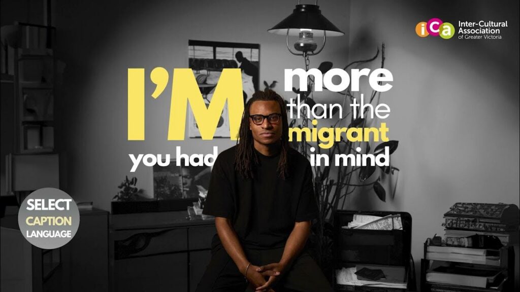 A man with dreadlocks and glasses sits looking forward, with text on screen reading "I'M more than the migrant you had in mind.