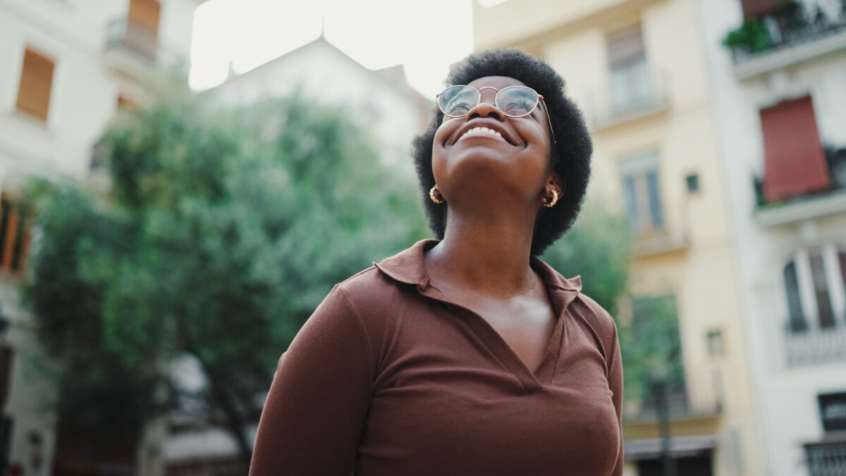 A smiling Black woman with glasses and a brown shirt looks up in front of buildings.
