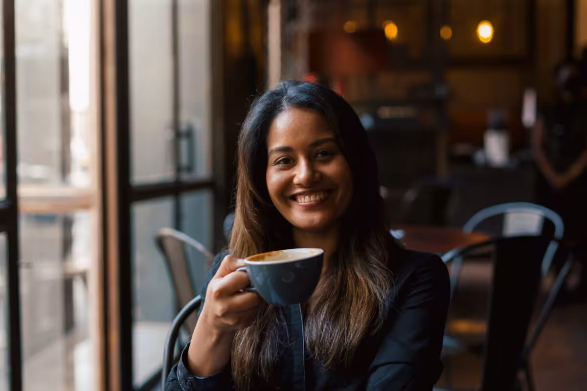 A smiling woman with long, dark hair holds a cup of coffee while sitting in a cafe.