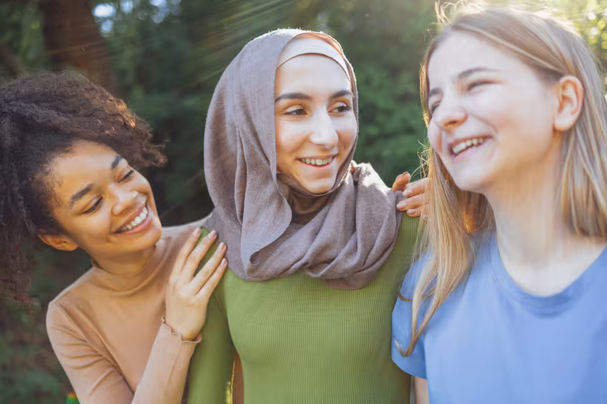 Three young women of diverse backgrounds smiling and laughing outdoors in dappled sunlight.