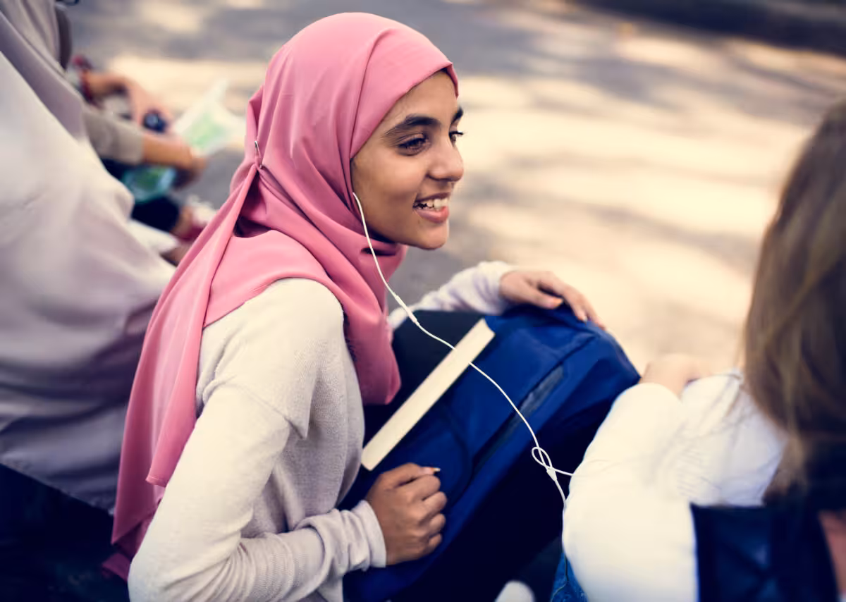 A young woman wearing a pink hijab smiles while listening to music on earphones.