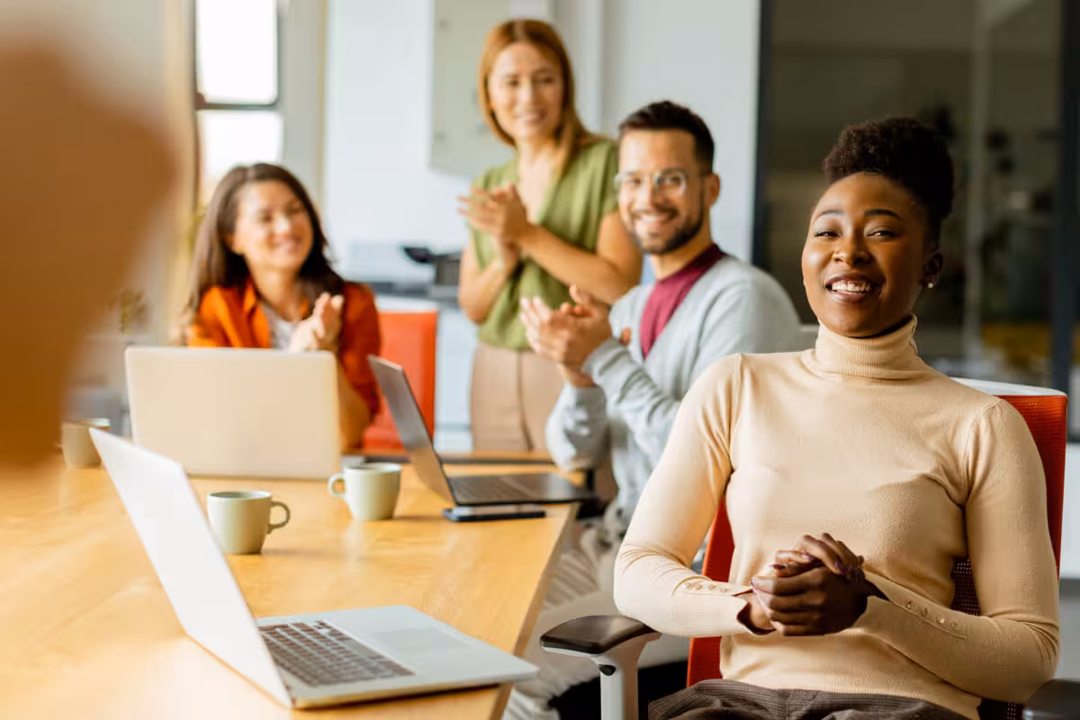 Diverse Group Colleagues Clapping Smiling During Meeting Around Diverse group of colleagues clapping and smiling during a meeting around a wooden table with laptops.