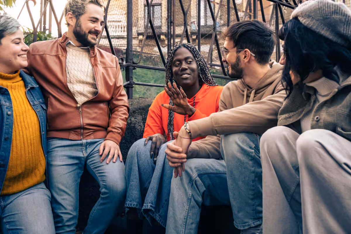 A diverse group of five young adults sit together, smiling and talking outdoors.