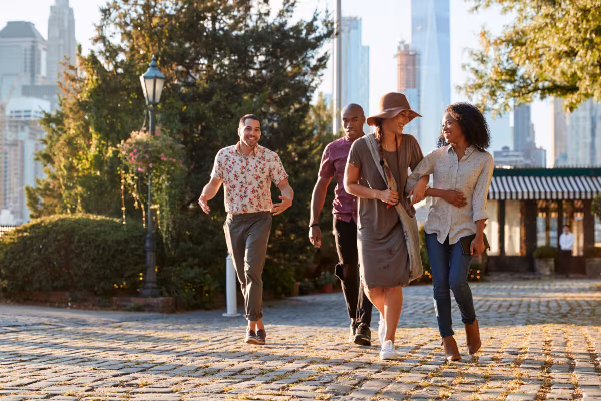 A diverse group of four friends walks along a cobblestone path in a city park, with buildings in the background.