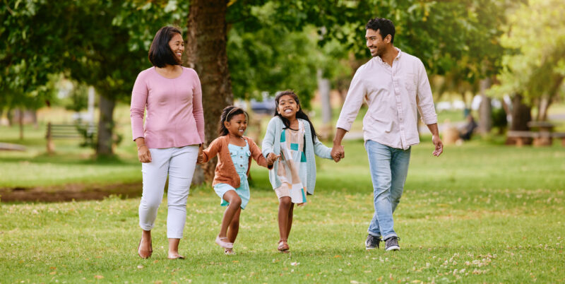 A family of four walks hand-in-hand across a grassy park on a sunny day.