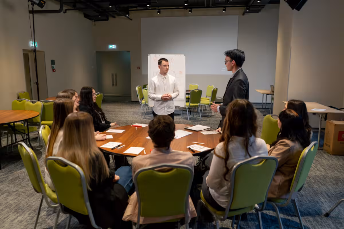 A group of people sit around a circular table in a conference room, listening to two men.