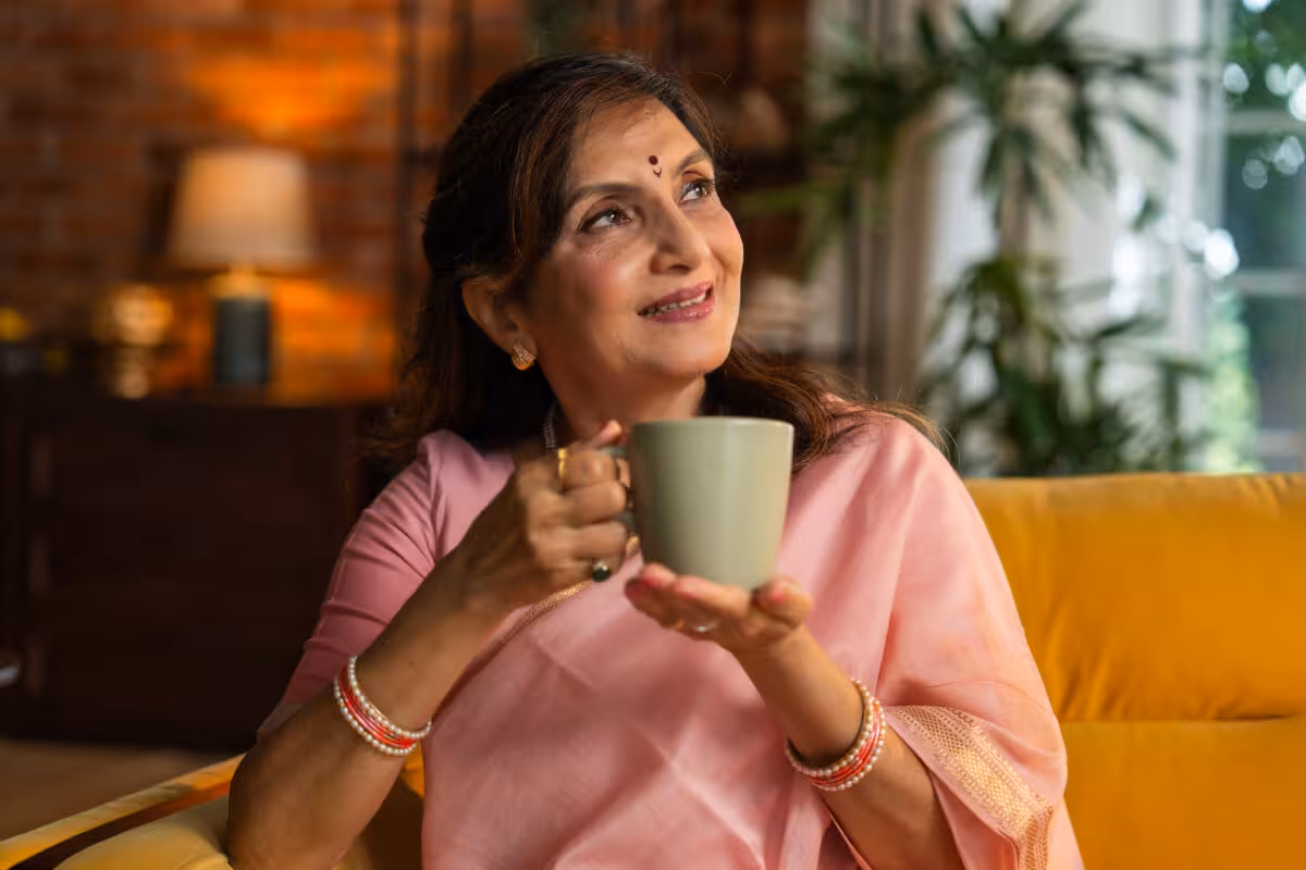 An Indian woman in a pink sari sits on a yellow couch, holding a mug and looking up and to the right.