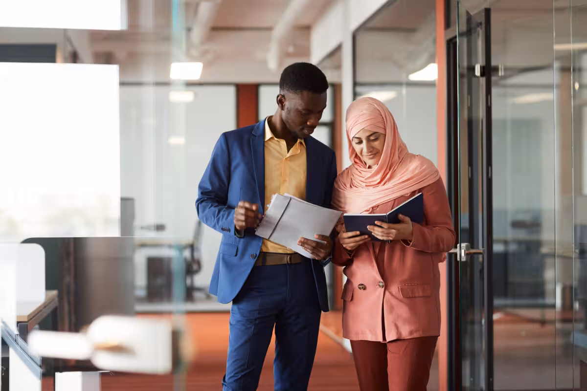 A man in a blue suit and a woman in a pink headscarf and suit look at documents together in an office hallway.