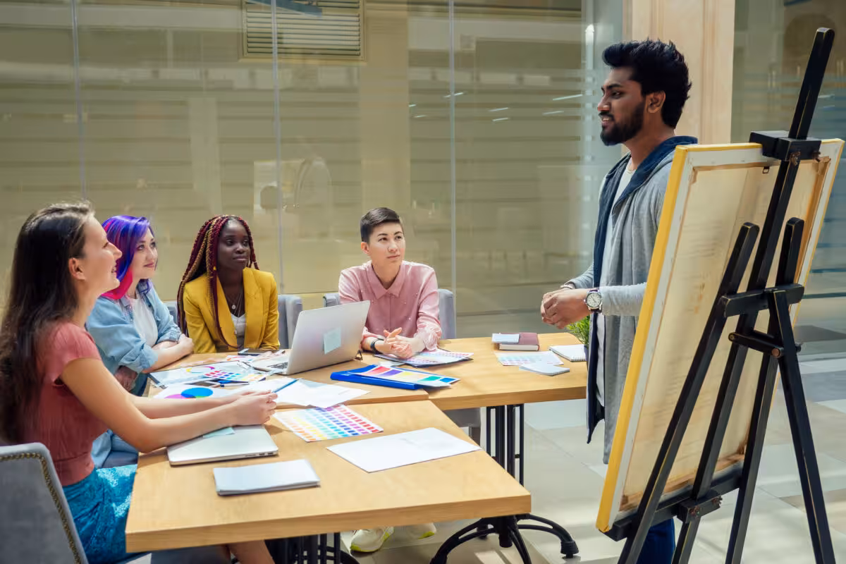 A man presents to a diverse group of people sitting at tables in a modern meeting room.