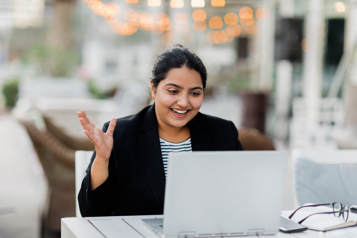 A smiling woman in a black blazer gestures while looking at a laptop on a table.