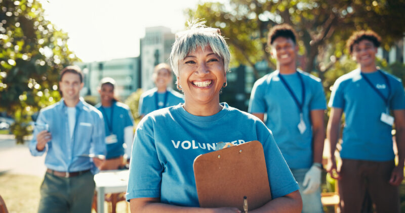 A smiling woman in a blue "VOLUNTEER" t-shirt holds a clipboard, with a diverse group of volunteers behind her.