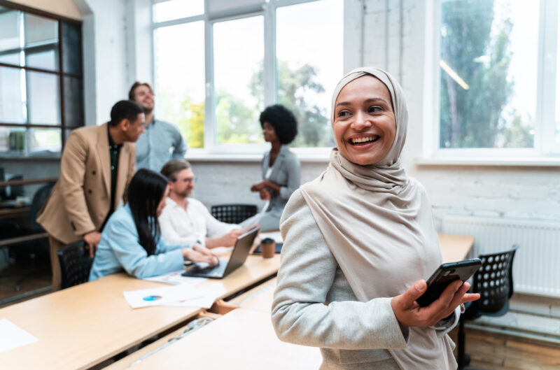 A smiling woman in a hijab holds a smartphone while a diverse group of colleagues works in the background.