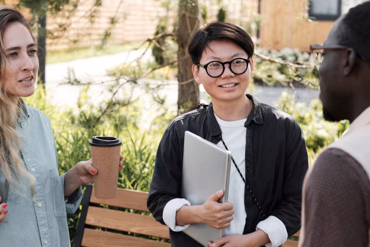 Three people talking outdoors, one holding a laptop and a coffee cup.