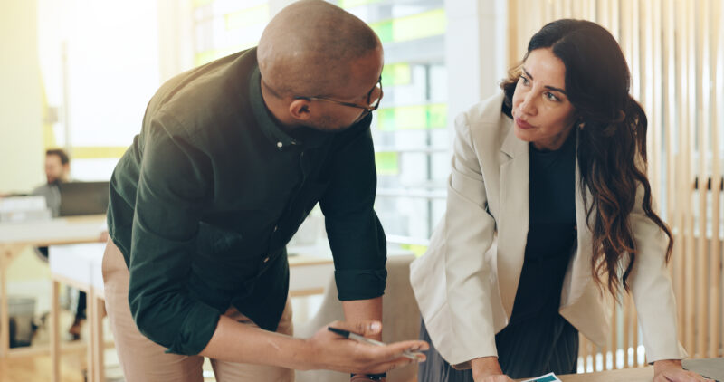 Two colleagues, a man and a woman, are looking at papers and discussing them at a table.