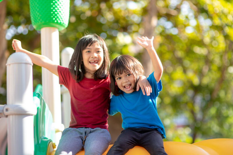 Two joyful children with arms around each other on a playground, with trees in the background.
