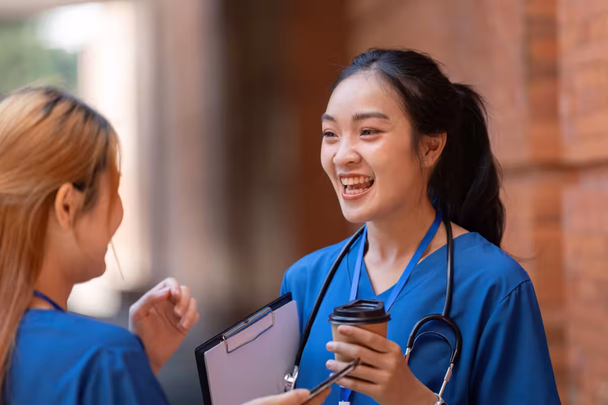 Two nurses in blue scrubs, one holding a coffee cup, laugh together outside a building.