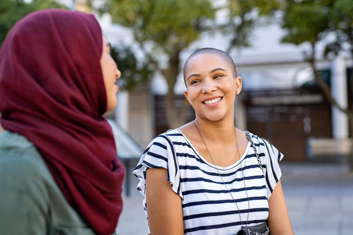 Two women, one wearing a hijab and the other with a shaved head, are talking outside.