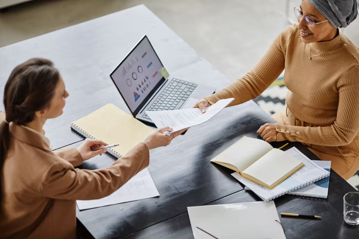 Two women at a table, one handing a document to the other, with a laptop and notebooks.